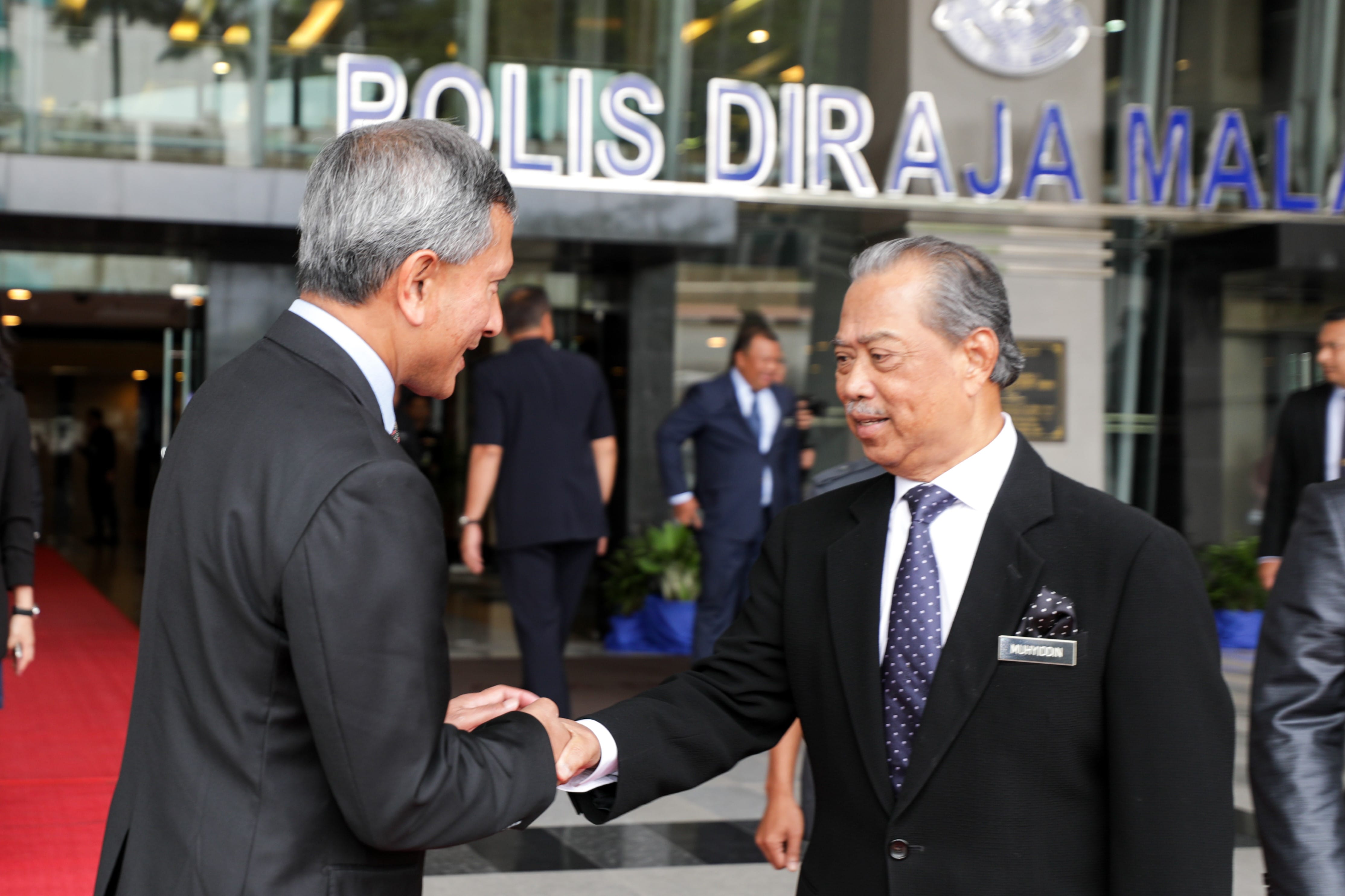 Two men in suits shaking hands outside a building with "Polis Diraja Malaysia" signage.